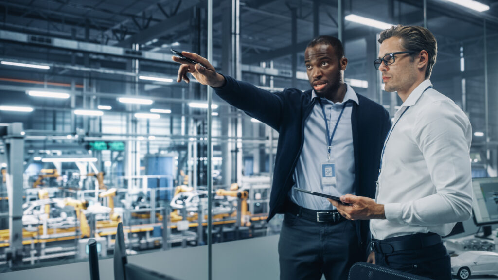 Two Diverse Automotive Industrial Engineers Talking About Vehicle Production while Standing in Office at a Car Assembly Plant. Industrial Specialists Discuss Work Projects on Tablet Computer.
