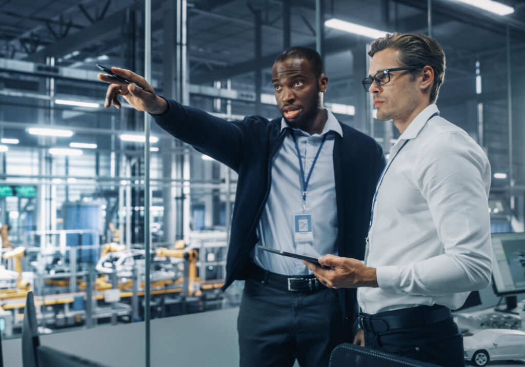 Two Diverse Automotive Industrial Engineers Talking About Vehicle Production while Standing in Office at a Car Assembly Plant. Industrial Specialists Discuss Work Projects on Tablet Computer.